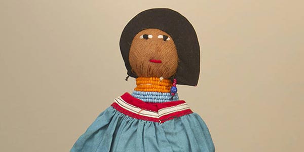 This is a coconut husk doll with a stump type body. Her facial features are stitched, and she wears a multicolored patchwork skirt and top, a beaded necklace, earrings, and a black bonnet.